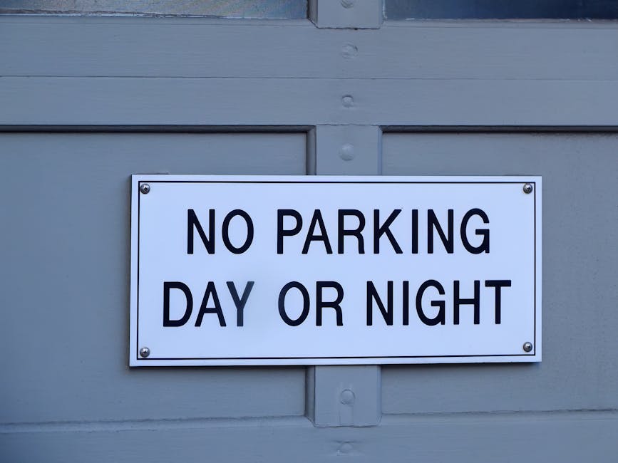 A rectangular white metal sign mounted on a light grey wooden exterior wall of a building, displaying the text 'NO PARKING DAY OR NIGHT' in bold black capital letters, secured with four screws at each