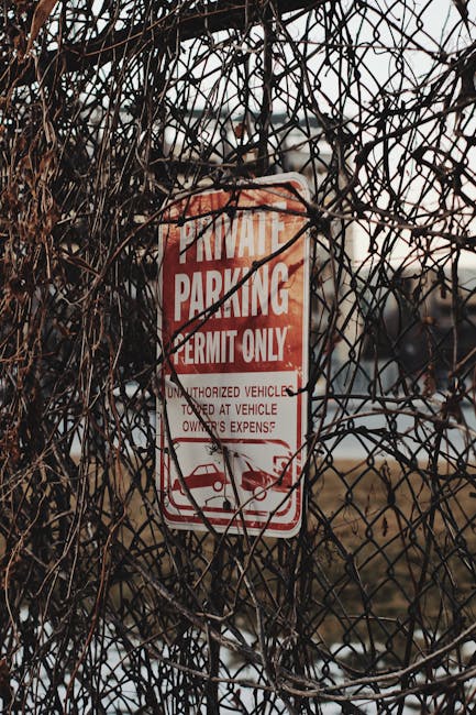A rectangular white metal sign mounted on a light grey wooden exterior wall of a building, displaying the text 'NO PARKING DAY OR NIGHT' in bold black capital letters, secured with four screws at each